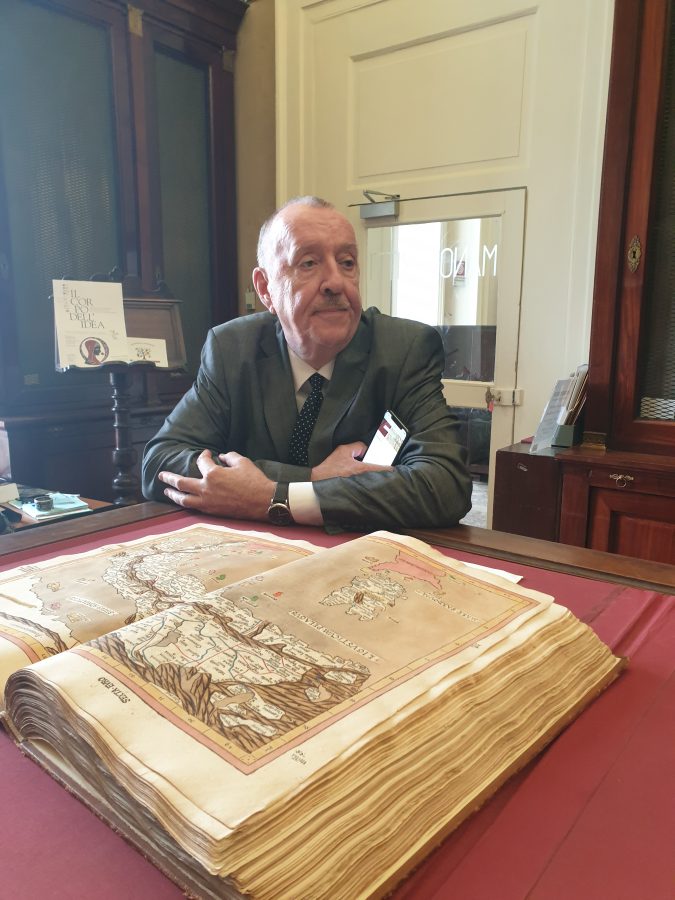 a man seated at a desk with his arms crossed and resting on the surface with a large manuscript atlas in the foreground