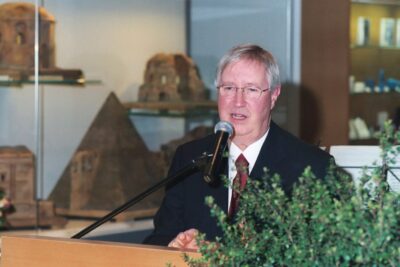 a photo of a white man with silver hair and glasses speaking at a microphone behind a podium.