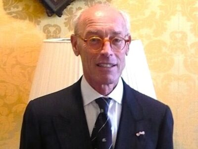 an older man in glasses wearing a dark suit in front of a yellow damask background